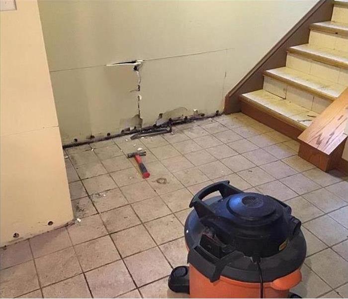 Dirty tile floor with white drywall with holes at the bottom with a stairway and an orange shop vac on the floor.